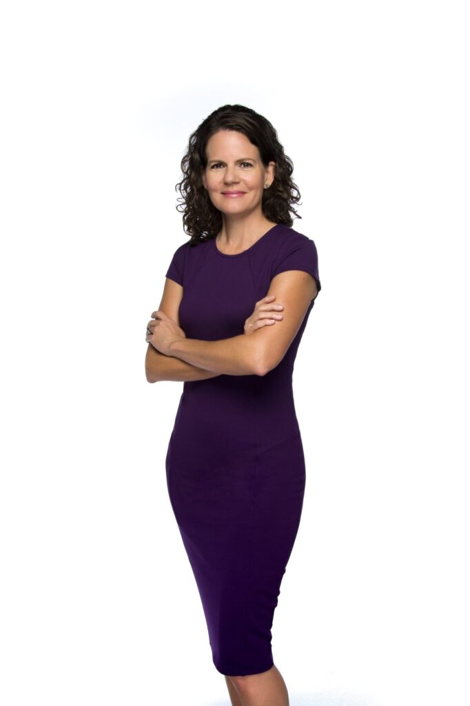 Woman in purple dress with wavy brown hair standing and smiling with her arms crossed